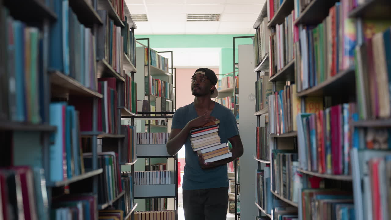 Student in casual t-shirt and cap holding large stack of books walks through library aisle, gazing thoughtfully at colorful shelves under bright indoor light