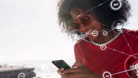 red de iconos digitales contra una mujer afroamericana que usa un teléfono inteligente en la playa