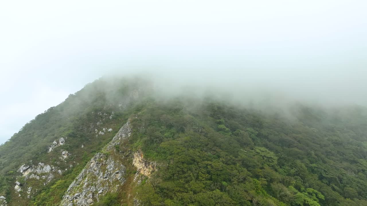 Pull‑out drone shot of dense fog sweeping over lush Cerro de Escamela above Orizaba, Mexico. Half frame green slope, half swirling sky—dramatic nature clip for travel, weather and cinematic use.