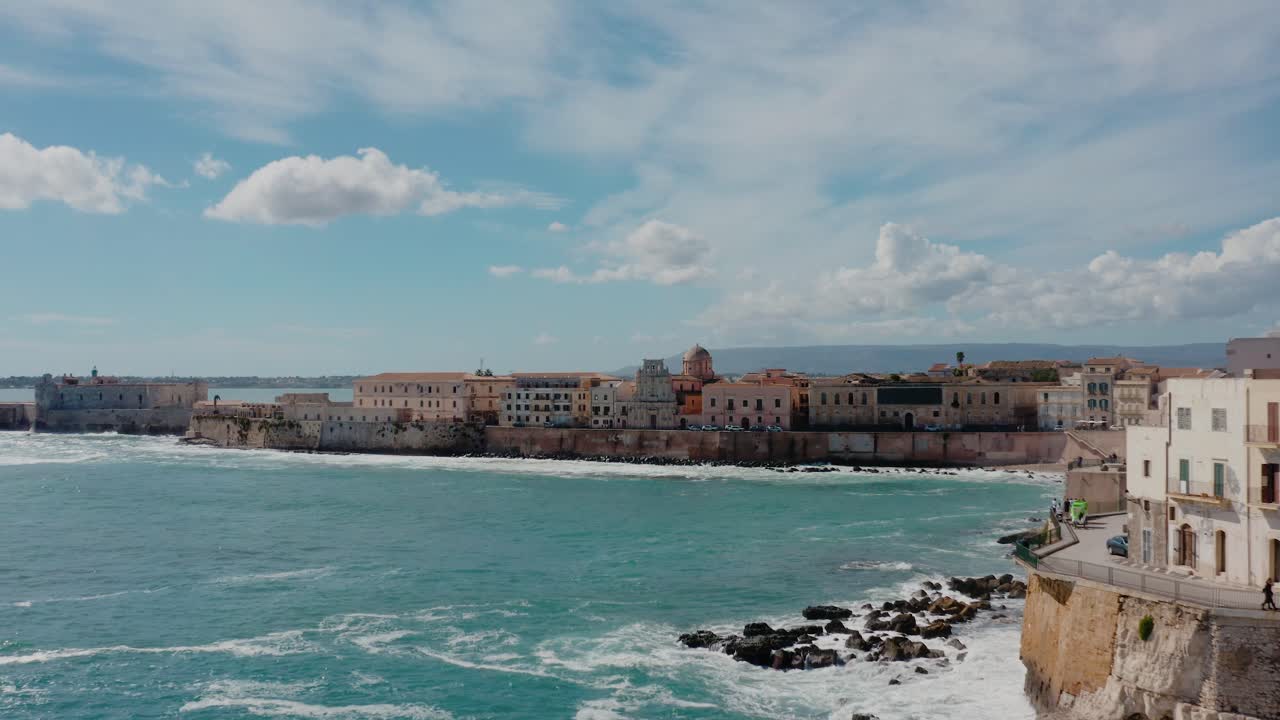 Flying along the cliff wall in Ortigia island with waves crashing on the shore. Aerial view of Ortigia island, Syracuse. Historic town in Sicily by Mediterranean sea.