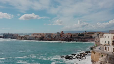 flying along the cliff wall in ortigia island with waves crashing on the shore. aerial view of ortigia island, syracuse. historic town in sicily by mediterranean sea.