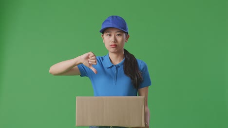 mensajera asiática insatisfecha en uniforme azul mostrando el pulgar hacia abajo mientras entrega una caja de cartón en el fondo de la pantalla verde en el estudio