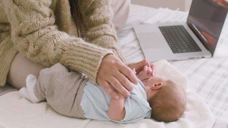 Unkenntliche Mutter arbeitet am Laptop und kuschelt ihr neugeborenes Baby, während sie zu Hause auf dem Bett sitzt