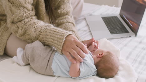 unrecognizable mother working on laptop computer and cuddling her newborn baby while sitting on the bed at home