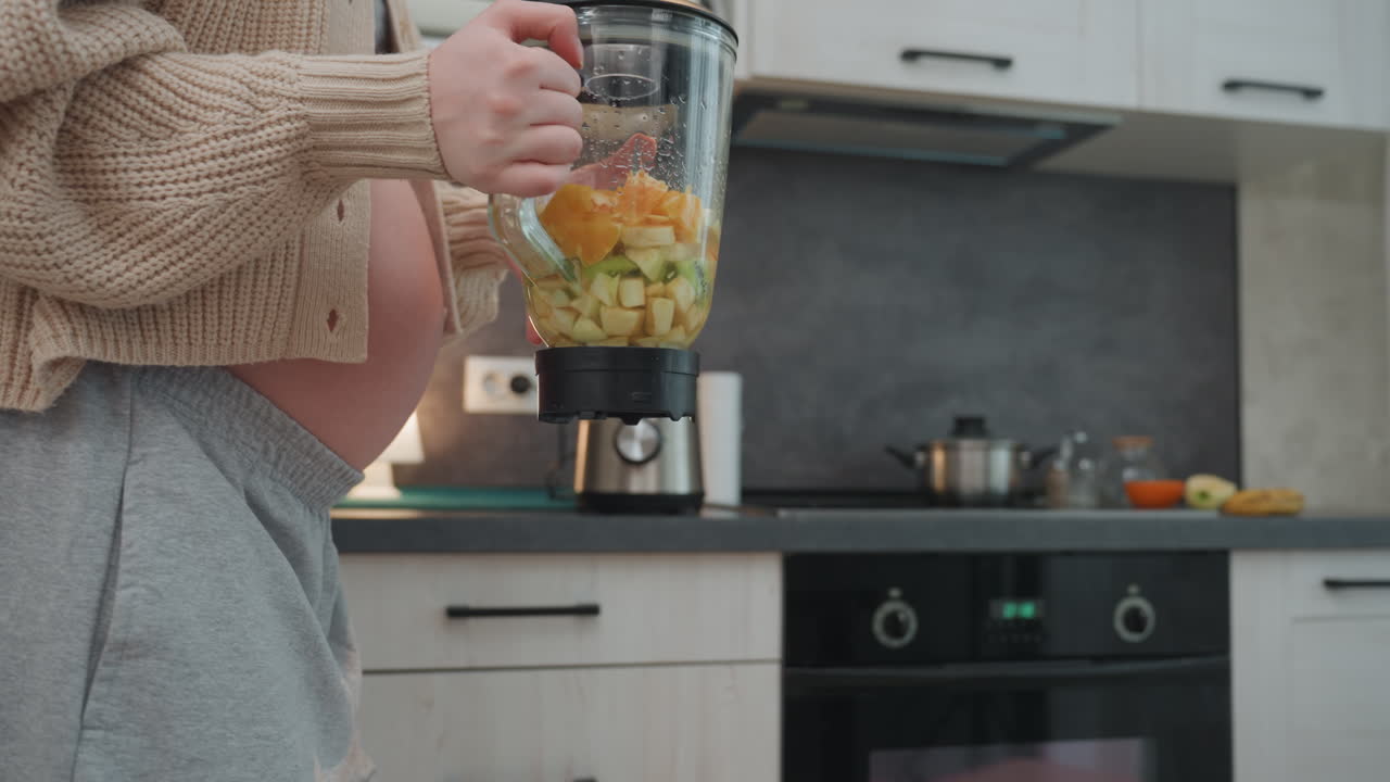 Donna bianca incinta che prepara un frullato di frutta, accogliente bancone della cucina, cardigan a maglia grossa, pancia visibile, che aggiunge mango e avocado al barattolo di vetro, che fissa il coperchio del frullatore, dolce routine mattutina, cuoca casalinga