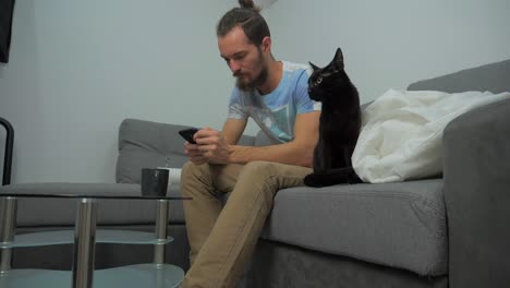 Young-man-is-checking-his-cell-phone-and-showing-the-screen-to-his-black-house-cat-sitting-on-the-couch