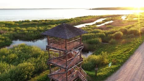 empty wooden lookout tower near lake and gravel road during golden sunset, aerial orbit view