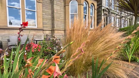 golden ornamental grass and vibrant flowers move in the wind outside a historic stone building, captured in bright daylight with a steady camera