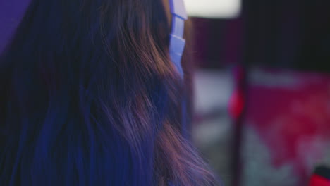 close up head view of woman with long brown hair wearing white headphones while playing game, vibrant lighting casting blue and red tones on hair with blurred colorful background in gaming room