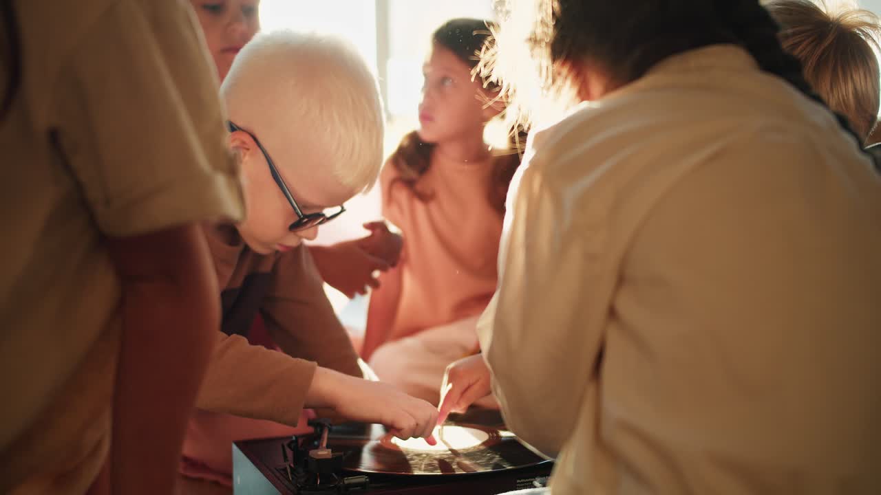 둥근 안경을 입은  머리카락을 가진 알비노 소년은 학교를 준비하는 클럽에서 친구들과 함께 레코드 플레이어를 조사하고 만져요.