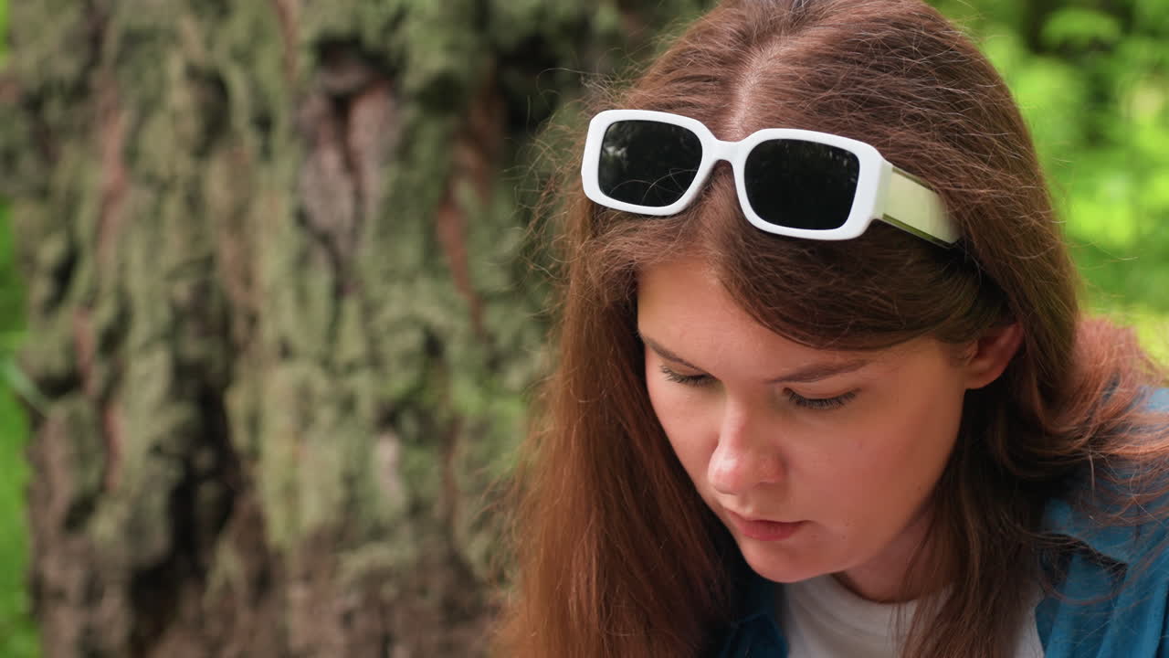 Close up of white woman wearing white sunglasses on head, focused on work outdoors, sunlight highlighting brown hair, calm expression, tree trunk behind, natural green background