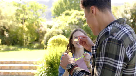 Feliz-Pareja-Diversa-Soplando-Burbujas-En-Un-Jardín-Soleado,-En-Cámara-Lenta