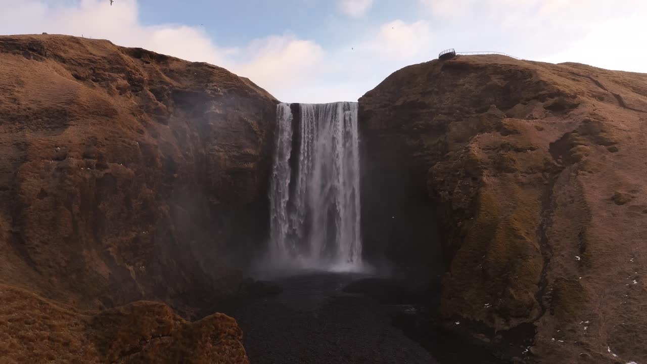 Drone slowly pushes toward Iceland’s iconic Skógafoss waterfall, mist, cliffs and golden highland terrain near Eyjafjallajökull volcano and Skógar valley.
