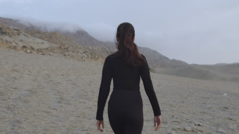 Slow-motion-medium-shot-from-the-back-of-a-woman-walking-through-a-desert-in-the-afternoon-with-cloudy-skies