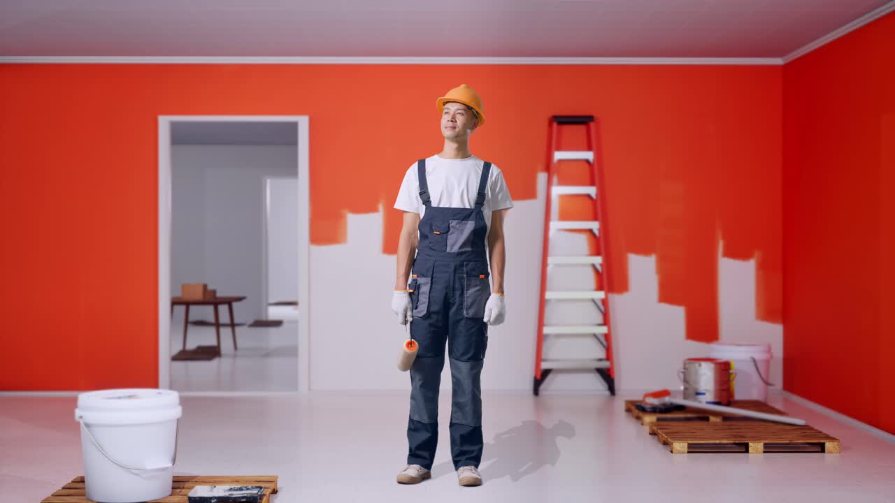 cuerpo lleno de pintor hombre asiático con casco de seguridad de pie y mirar a su alrededor con la pared de pintura en la habitación