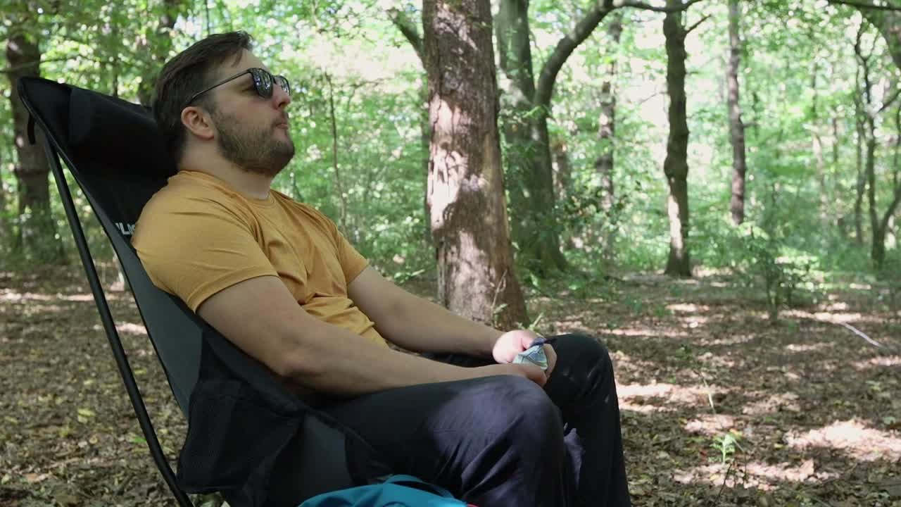 hombre con gafas de sol come un bocadillo y descansa sentado en una silla en un campamento en el bosque