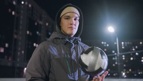 jovem adulto vestindo capuz segurando uma bola de futebol na mão e olhando com confiança para a câmera durante a noite em um campo esportivo urbano com edifícios residenciais iluminados e luzes de rua