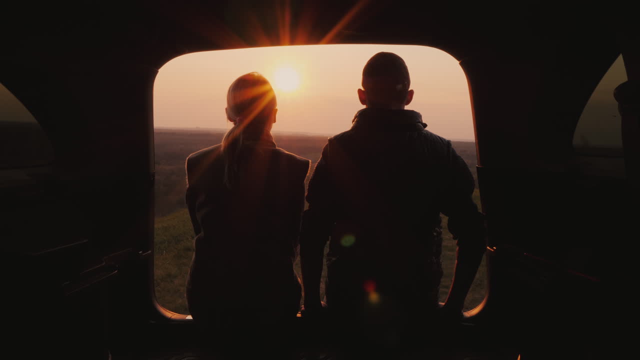 한 젊은 부부가 그림 같은 곳에서 아름다운 일몰을 감상하며 차 트렁크 옆에 앉아 있다