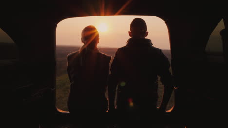 a young couple sits next to the trunk of a car admiring the beautiful sunset in a picturesque place