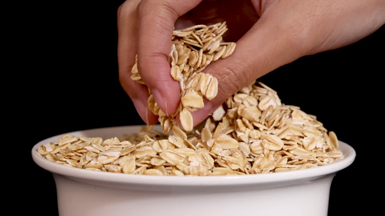 A hand sifts through oats in a bowl, highlighting texture and movement against a black background with soft lighting
