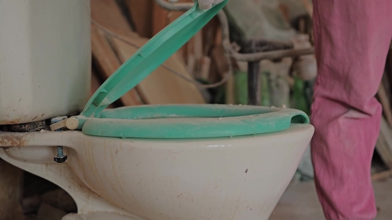 Slow motion captures gloved hand lifting dusty toilet lid inside renovation site