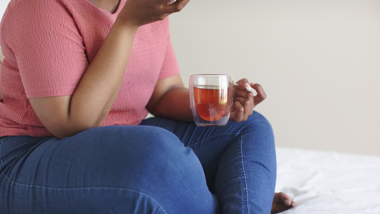 Mulher atendendo uma ligação ao levantar o smartphone, falando ao telefone e tomando um gole de chá no sofá