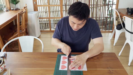 asian man cutting heart shape paper using utility knife cutter with ruler on cutting mat