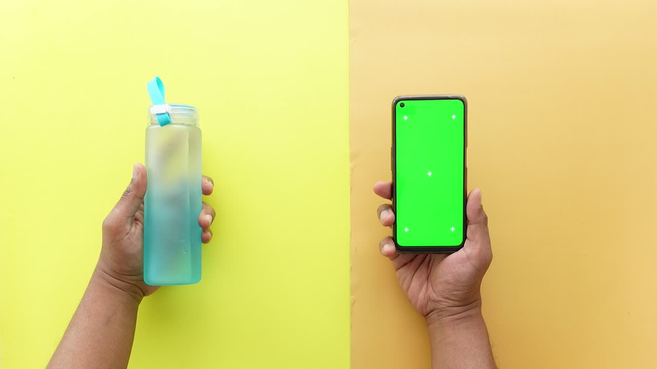 Hands holding water bottle and smartphone with green screen on a yellow background