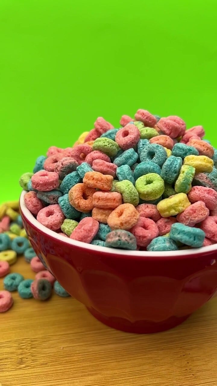 Bunte ringförmige Cerealien füllen eine weiße Schüssel, mit rosa, blauen, grünen, gelben und orangefarbenen Stücken vor einem leuchtend grünen Hintergrund in Nahaufnahme.