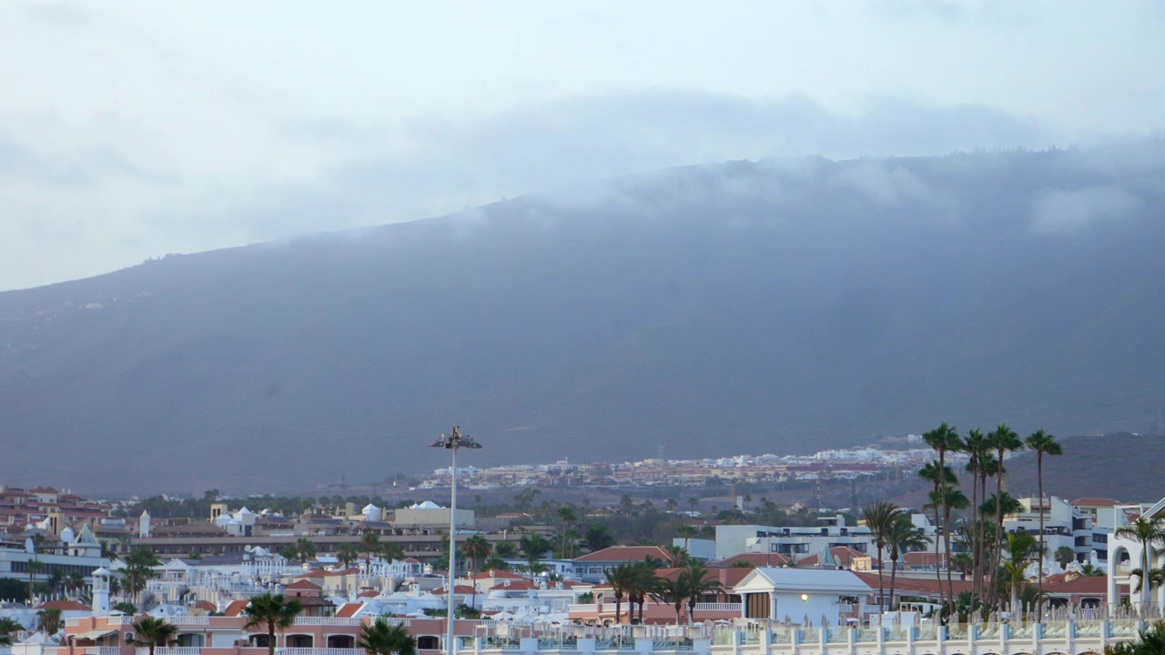 Vuoret nousevat matalien pilvien läpi valkoisten lomarakennusten ja palmujen yläpuolella Costa Adejessa Teneriffalla. Laaja iltanäkymä vangitsee utuisen vulkaanisen maiseman.