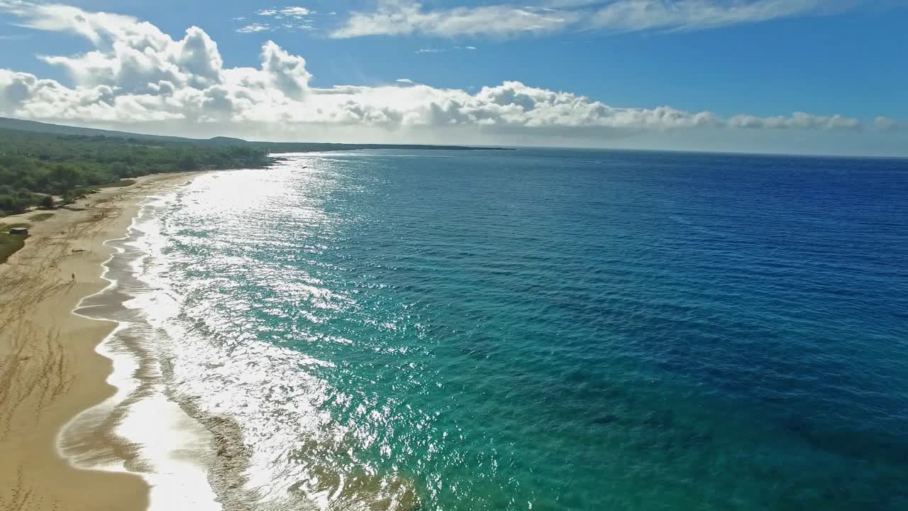 Toma aérea panorámica hacia el sol con una persona caminando por la costa de Maui, Hawái, con hermosas aguas azules y montañas.