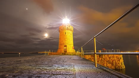 vista de ángulo bajo del faro iluminando por la noche con la luna y las nubes en el fondo