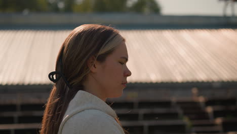 young adult with long hair tied back, wearing hoodie, walks thoughtfully by empty stadium bleachers on sunny day, she appears reflective and deep in thought