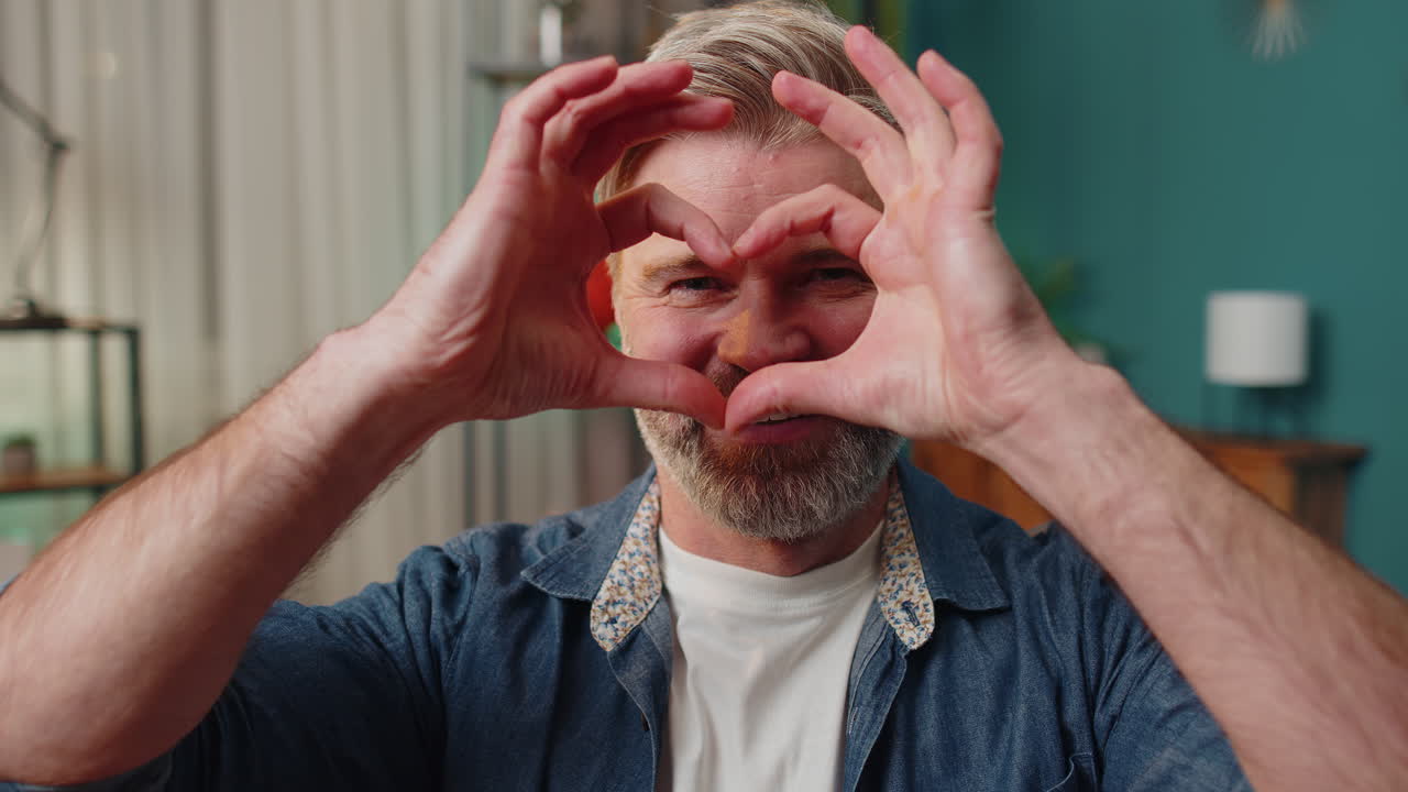 un hombre feliz de mediana edad hace un símbolo de amor mostrando un signo de corazón a la cámara expresando sentimientos románticos