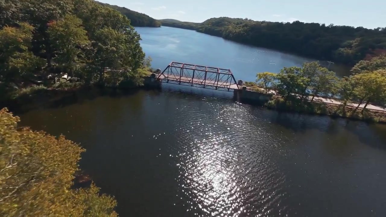 Aerial view of Muscoot Reservoir in Westchester, NY. Historic rusty bridge over the water surrounded by trees on a sunny day. The camera dolly in over trees and boom down to the bridge. Dynamic moves.