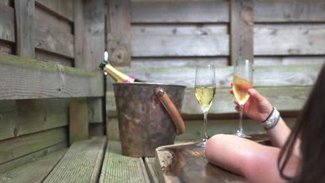 couple sat in luxury cedar wood hot tub grabbing a glass of champagne sat on the side next to a champagne bottle in ice box