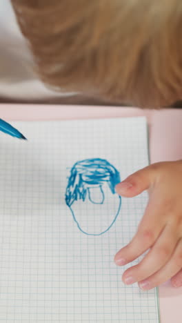 blond little boy colors doughnut with blue marker on exercise book page at table closeup. elementary school student draws picture at art lesson