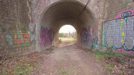 Walking-through-a-tunnel-with-light-at-the-end,-sun-beams-and-flares-on-street-art-and-graffiti-on-the-stone-walls