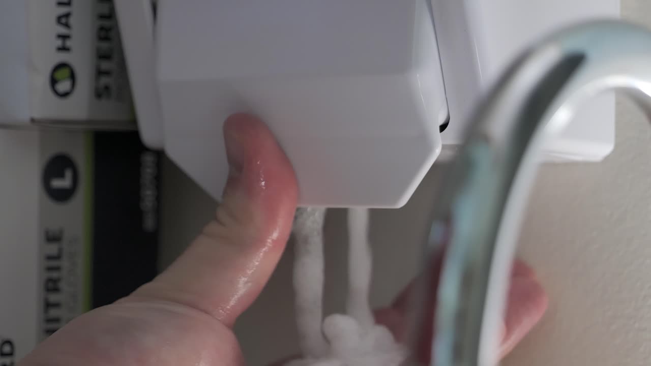 A person is seen squirting soap onto their hand. Medical gloves can be seen nearby.