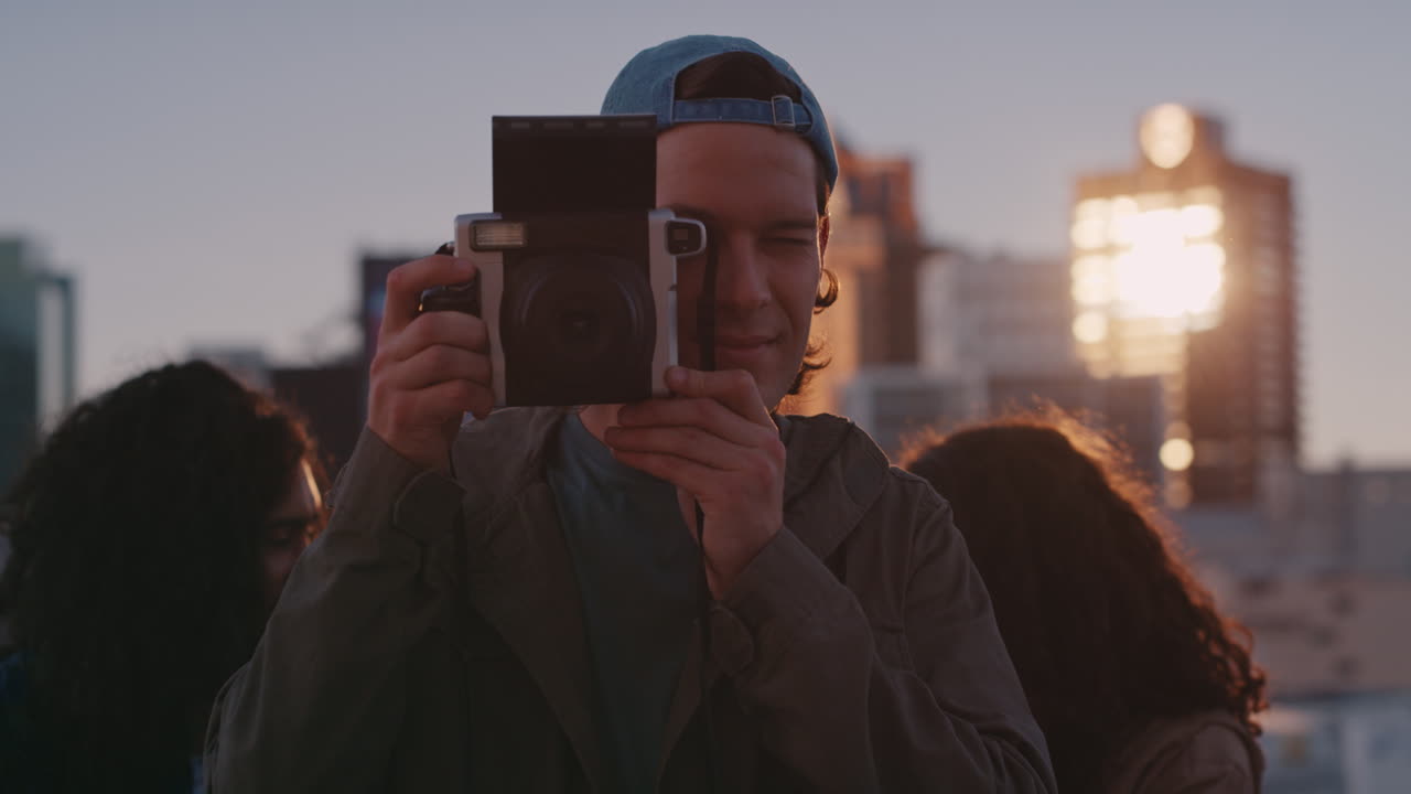 happy friends hanging out on rooftop young man using polaroid camera photographing weekend party celebration at sunset