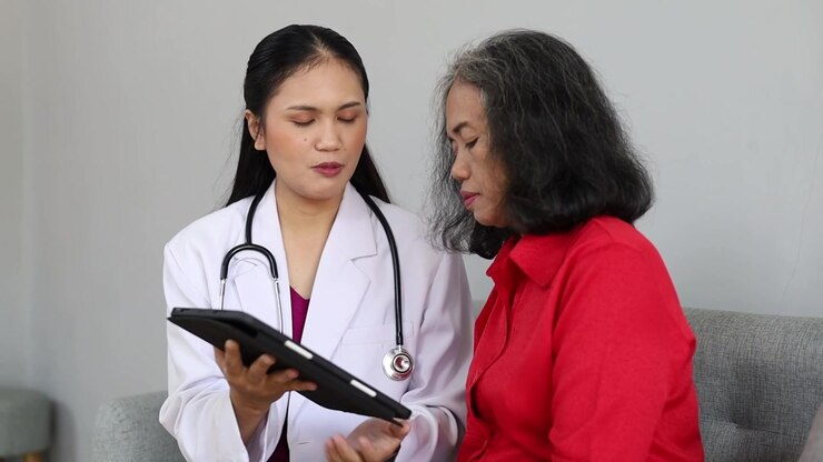Female doctor using a digital tablet to explain medical information to an elderly Asian woman during a home healthcare consultation.
