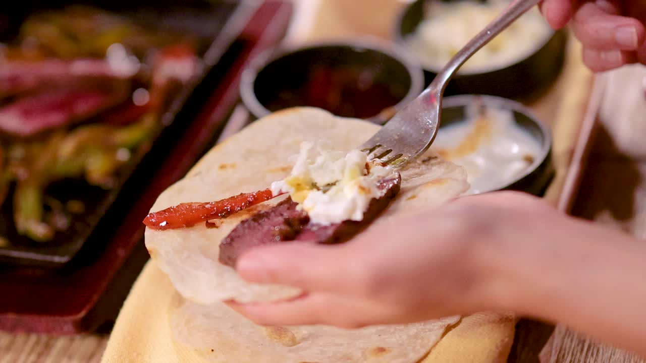 Hand adds cheese to beef taco with salsa and sour cream, warm indoor lighting, close-up