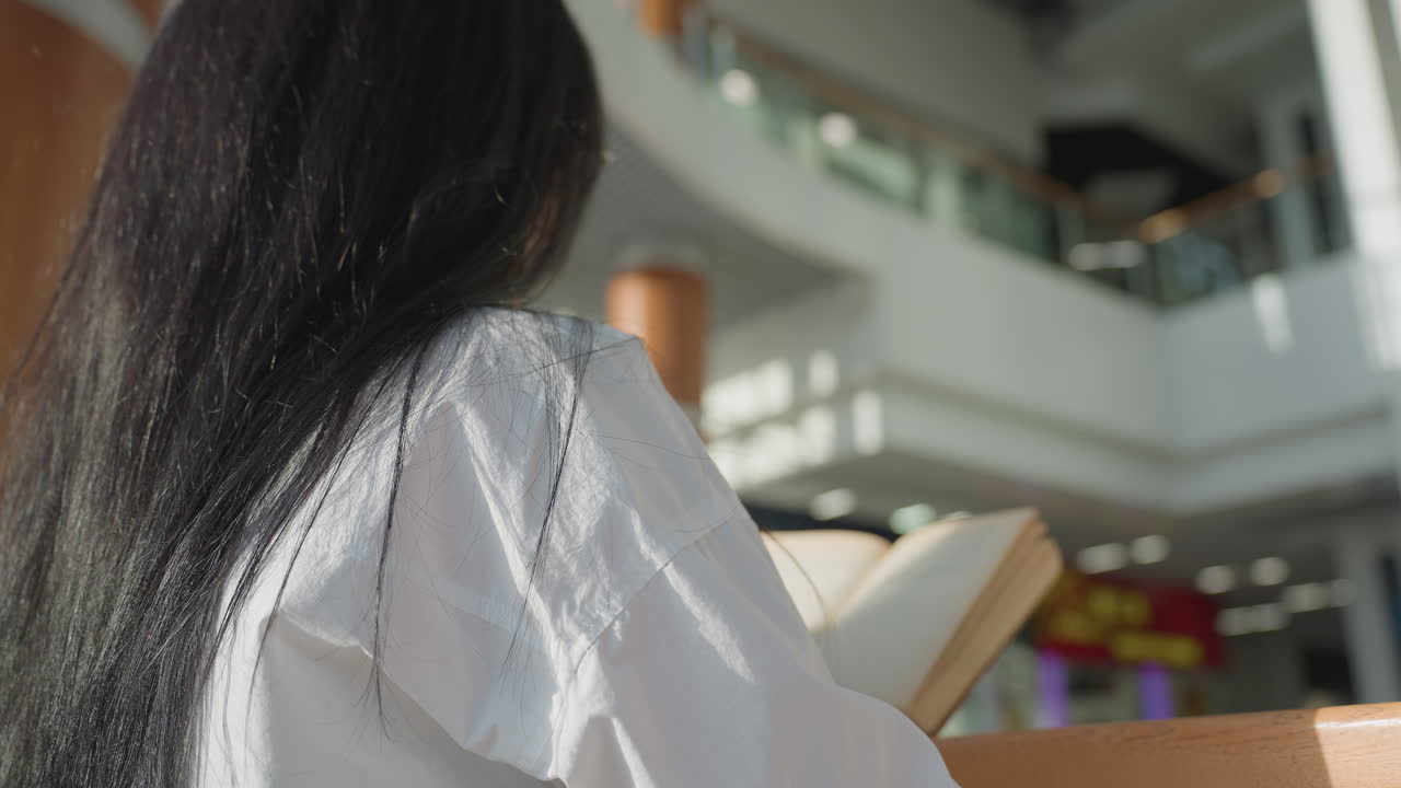 Behind view of woman with long black hair reading open book while standing by wooden rail in brightly lit indoor space, colorful reflections blinking softly in background