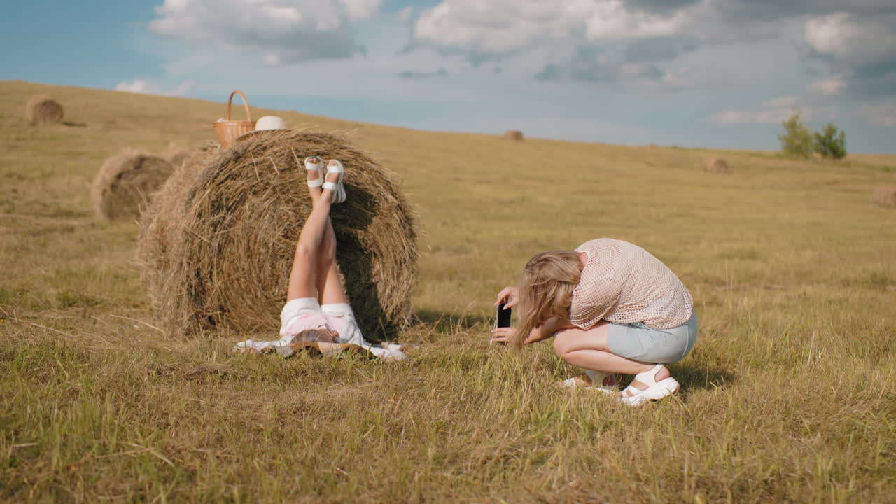 задний вид женщины, фотографирующей друга, лежащего на сено с поднятыми ногами, наслаждающегося солнечным сельским пейзажем, корзиной для пикника и шляпой, отдыхающей на сене, в то время как золотое поле простирается на расстояние под огромным небом