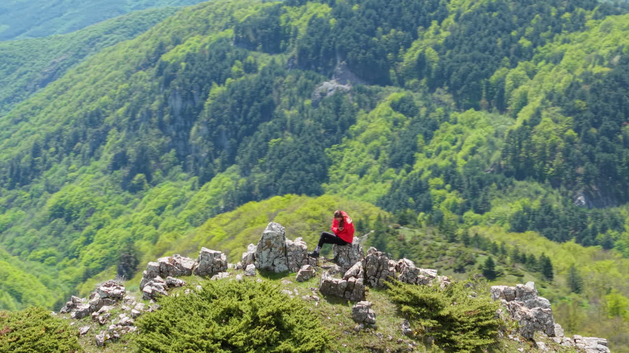 Drone shot of a traveler standing confidently on a mountain summit in Beklemeto Pass, Bulgaria, surrounded by lush green valleys and forested hills. A tranquil moment of connection with nature.