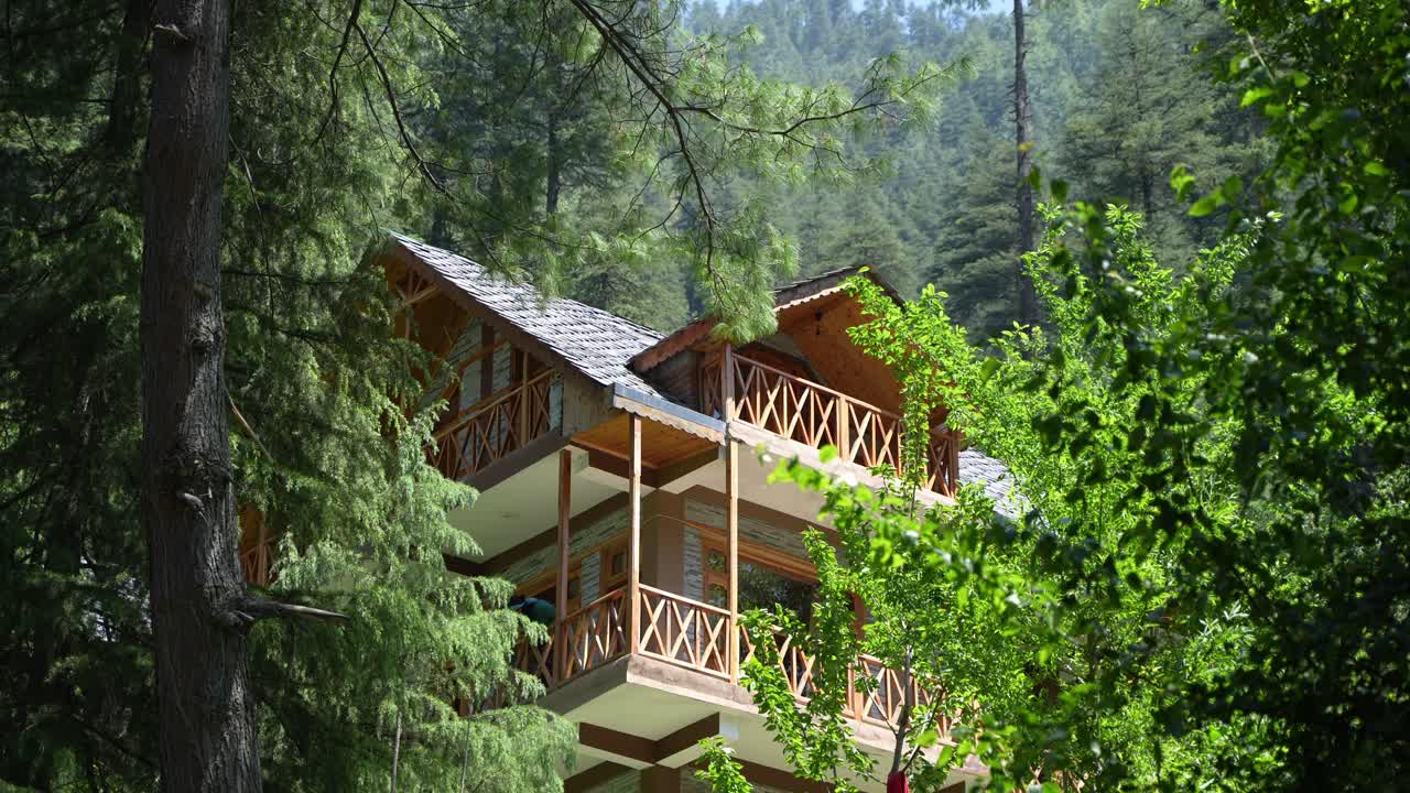 A wooden house is seen through a gap in the trees in a mountainous area.