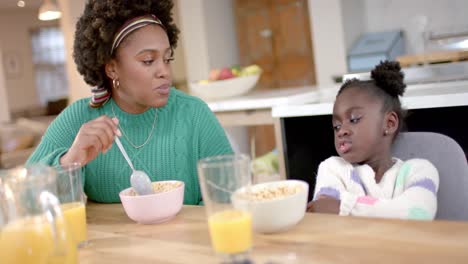 African-american-mother-and-daughter-eating-cereal-with-milk-and-talking-in-kitchen,-slow-motion