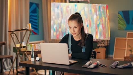 un hermoso y joven pintor trabaja en un salón, un estudio de pintura, se sienta en el escritorio frente a un monitor de portátil, hace clic en el teclado, envía correos electrónicos, mira a la cámara, cruza los brazos con una sonrisa