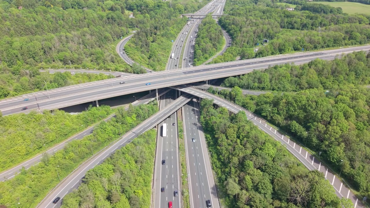 Perspektif udara menunjukkan persimpangan jalan tol yang kompleks. Beberapa lajur lalu lintas mengalir di jalan-jalan yang berkelok melalui pedesaan hijau yang luas