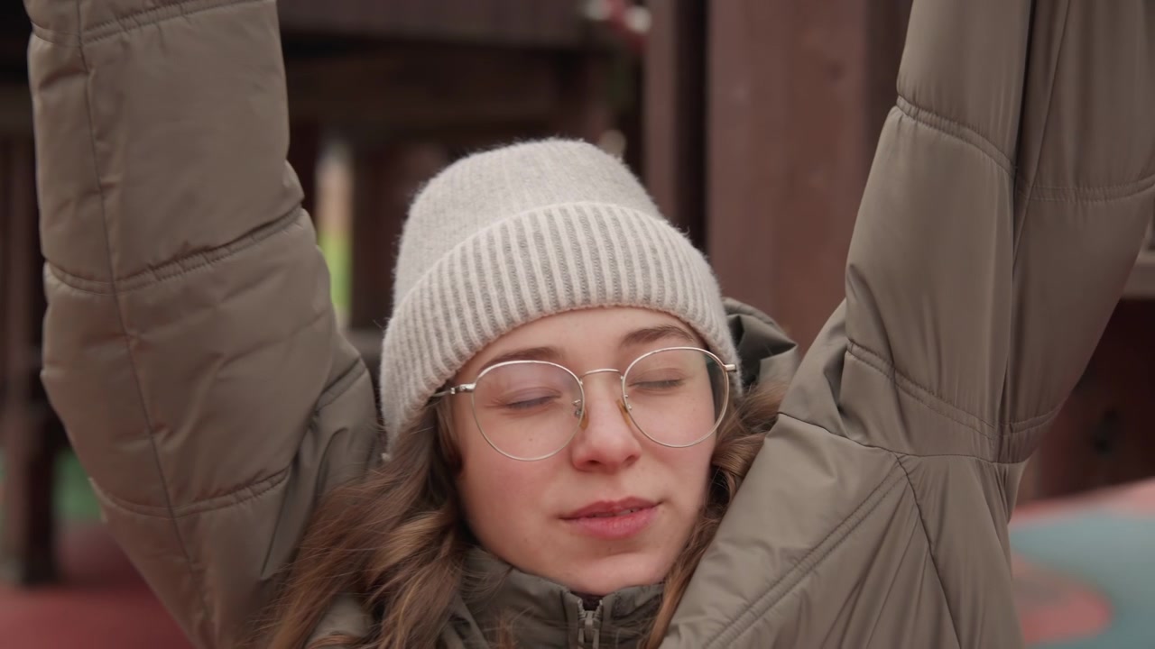 White woman resting on playground rings with relaxed smile, wearing knitted beanie and glasses, winter coat folded arms, closed eyes and calm breath, park background with soft trees and warm muted